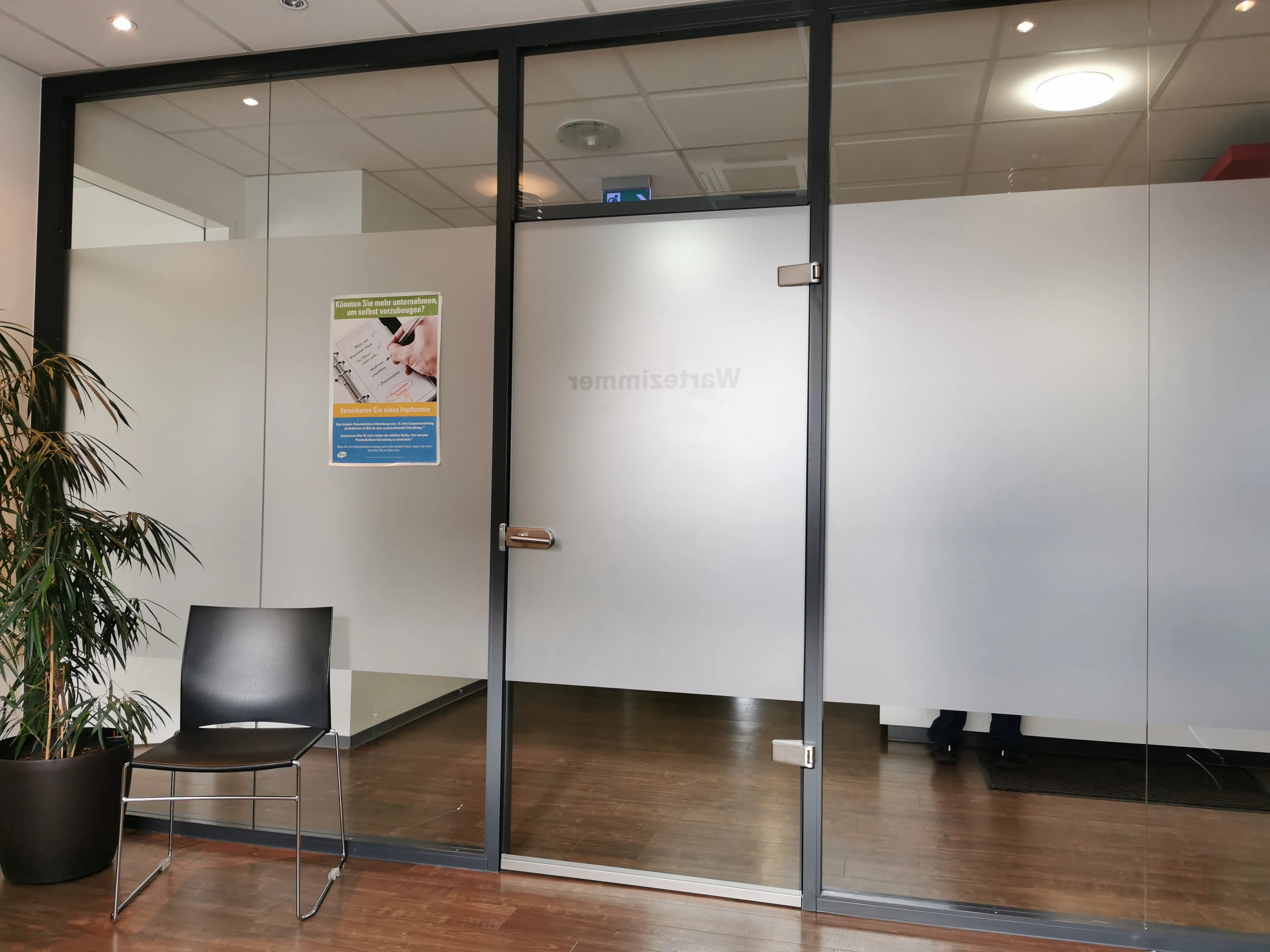 Bir Alman doktor muayenehanesinde üzerinde 'Wartezimmer' (bekleme odası) yazan cam kapı girişi, önünde bir sandalye ve saksı bitkisi
