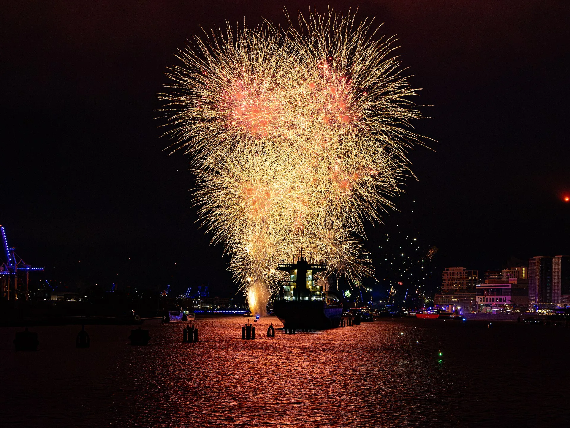 Hamburg limanında Silvester gecesi bir gemi üzerinde patlayan altın rengi havai fişekler ve suya yansıyan ışıklar