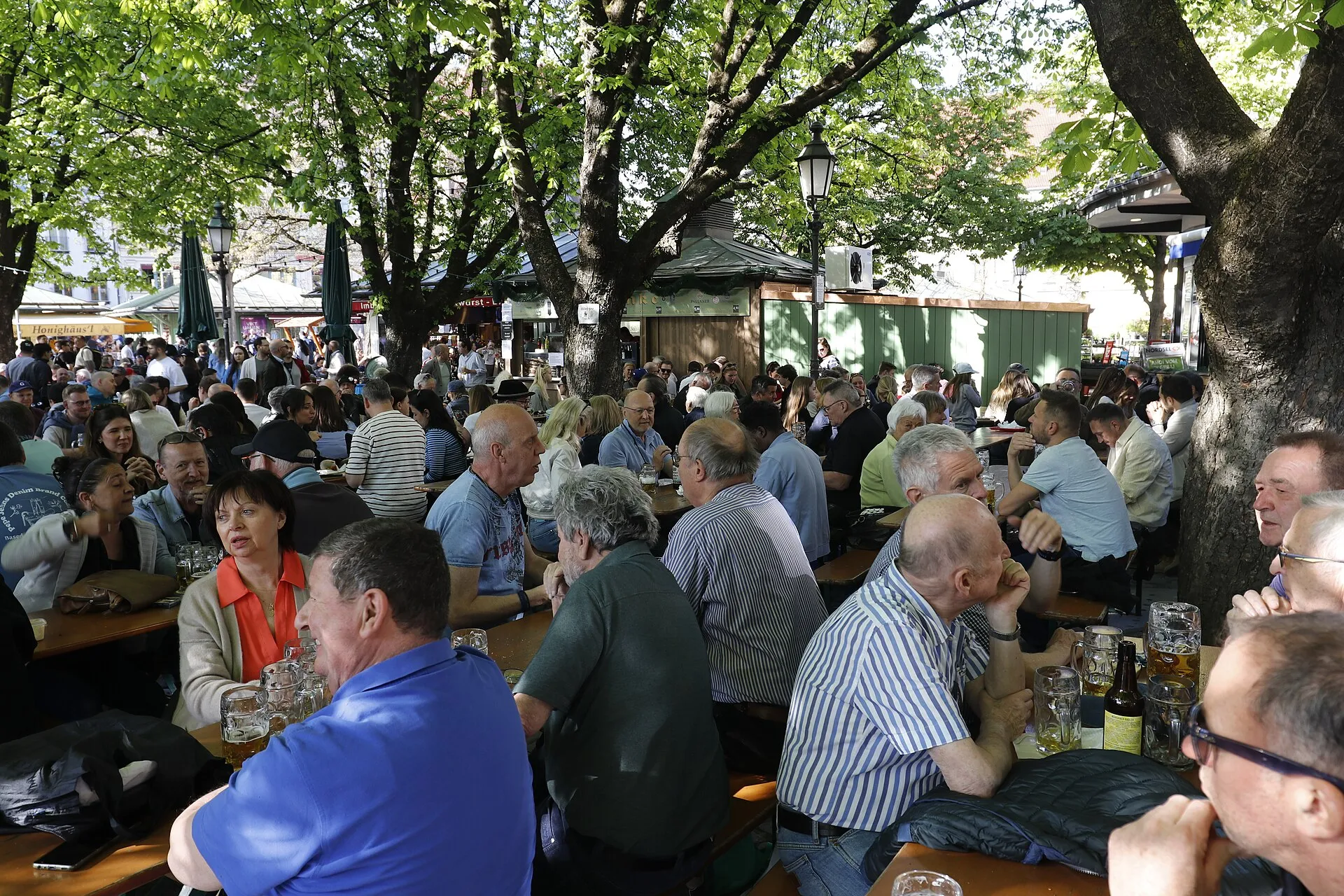 Münih Viktualienmarkt'taki gölgeli kestane ağaçları altında kalabalık masalarda bira içen ziyaretçilerin bulunduğu geleneksel bir Biergarten