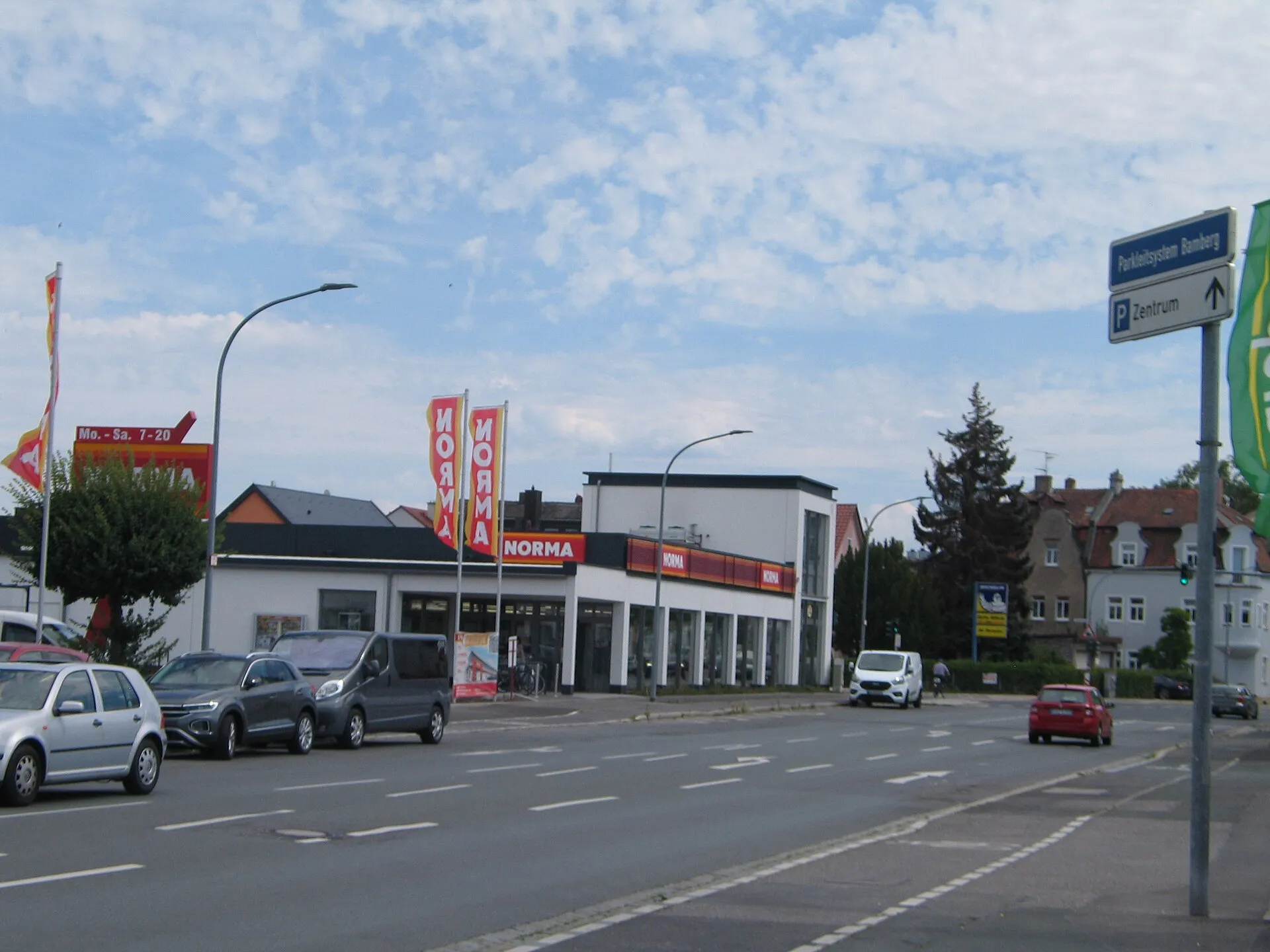 Bamberg'de bir Norma discounter marketinin dışından görünümü - kırmızı-sarı NORMA tabelaları ve 'Mo.-Sa. 7-20' açılış saatleri yazan pankart