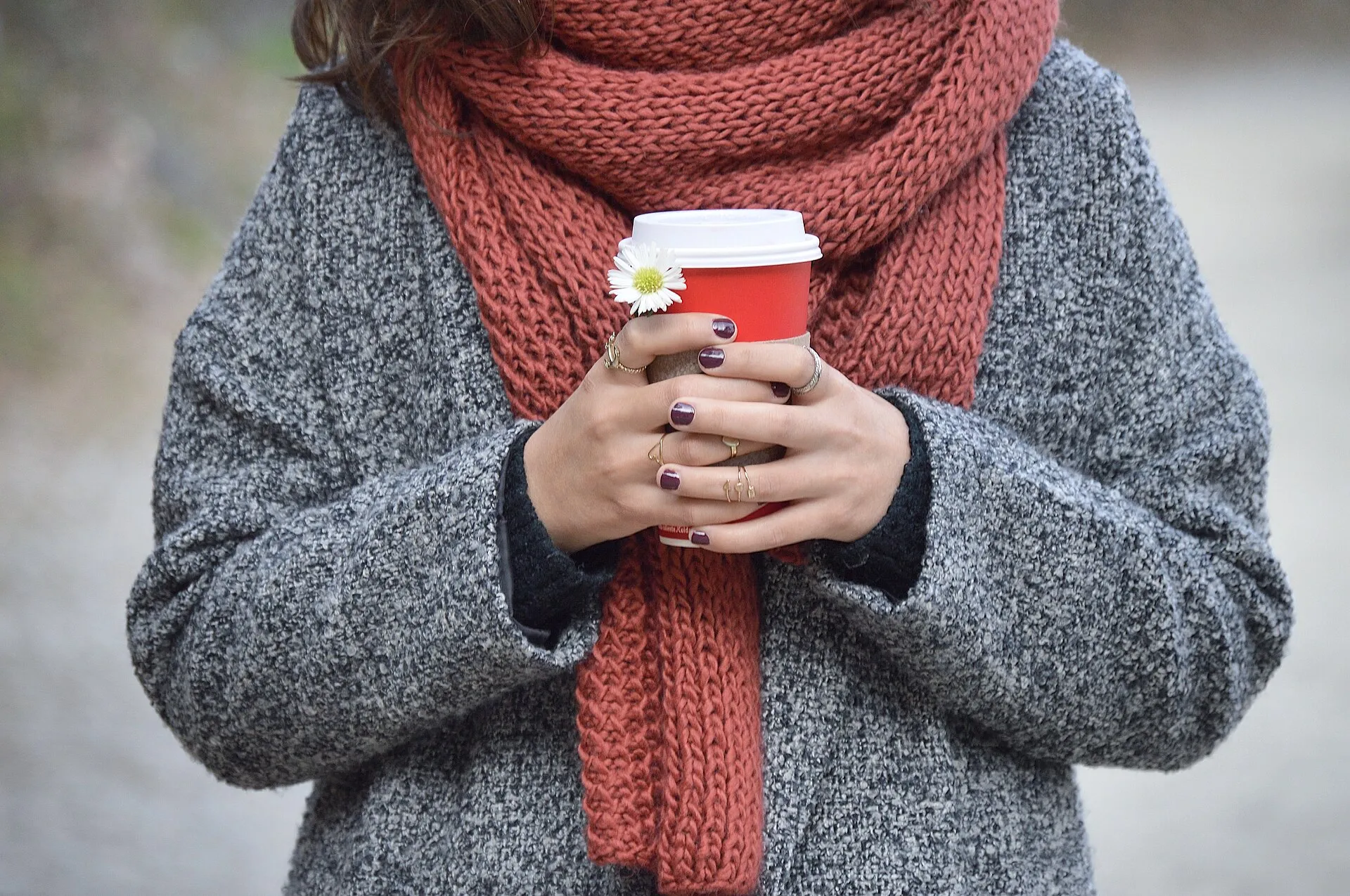 Örgü atkı ve kalın kazak giyen bir kişinin elinde kırmızı, kapaklı tek kullanımlık kahve bardağı (Kaffee zum Mitnehmen) tutması