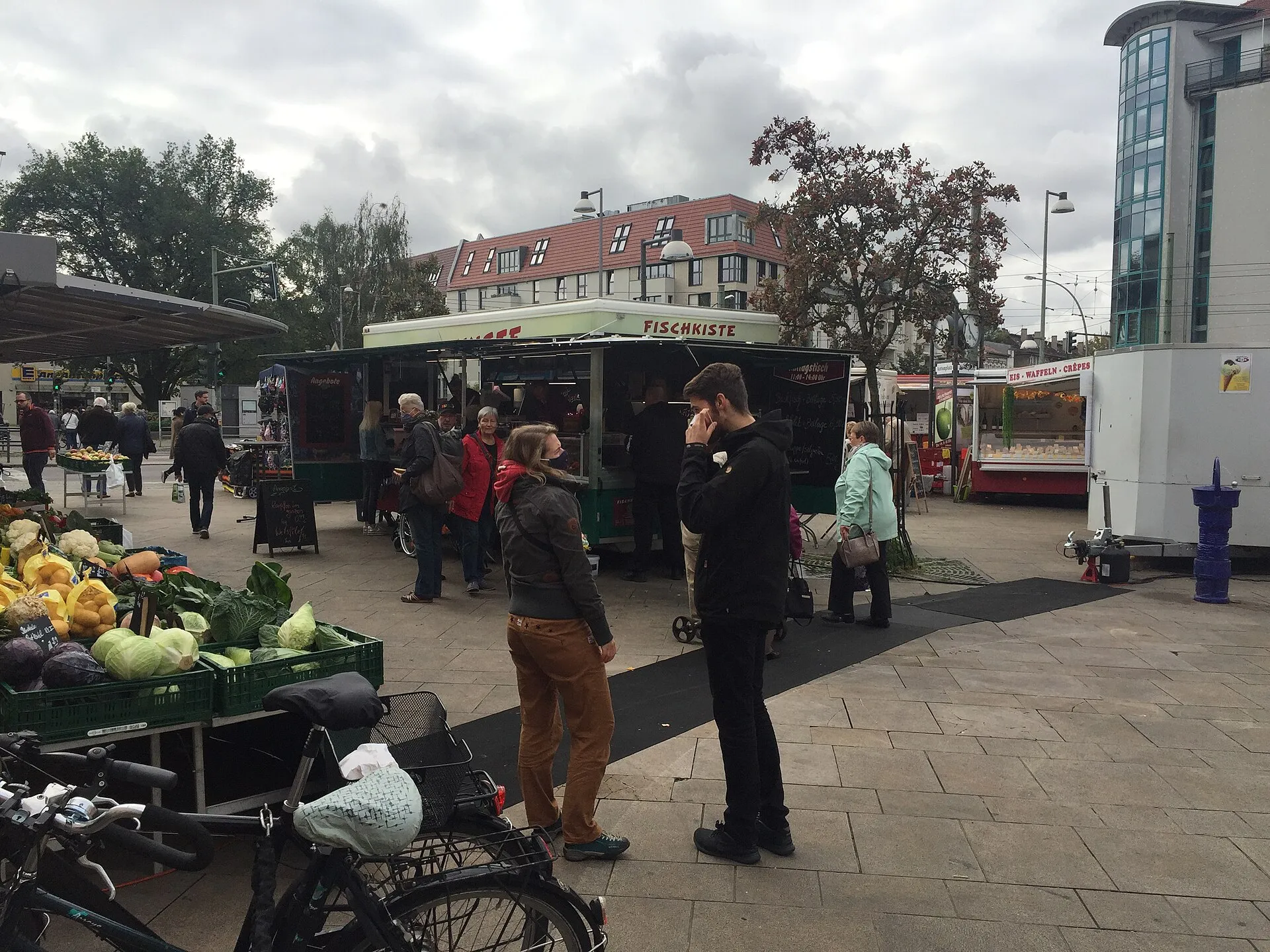 Berlin'de bir Wochenmarkt (haftalık semt pazarı) sahnesi - kasalar içinde taze lahana, karnabahar ve limonlar, arka planda bir balık satan Fischkiste tezgahı ve alışveriş yapan insanlar