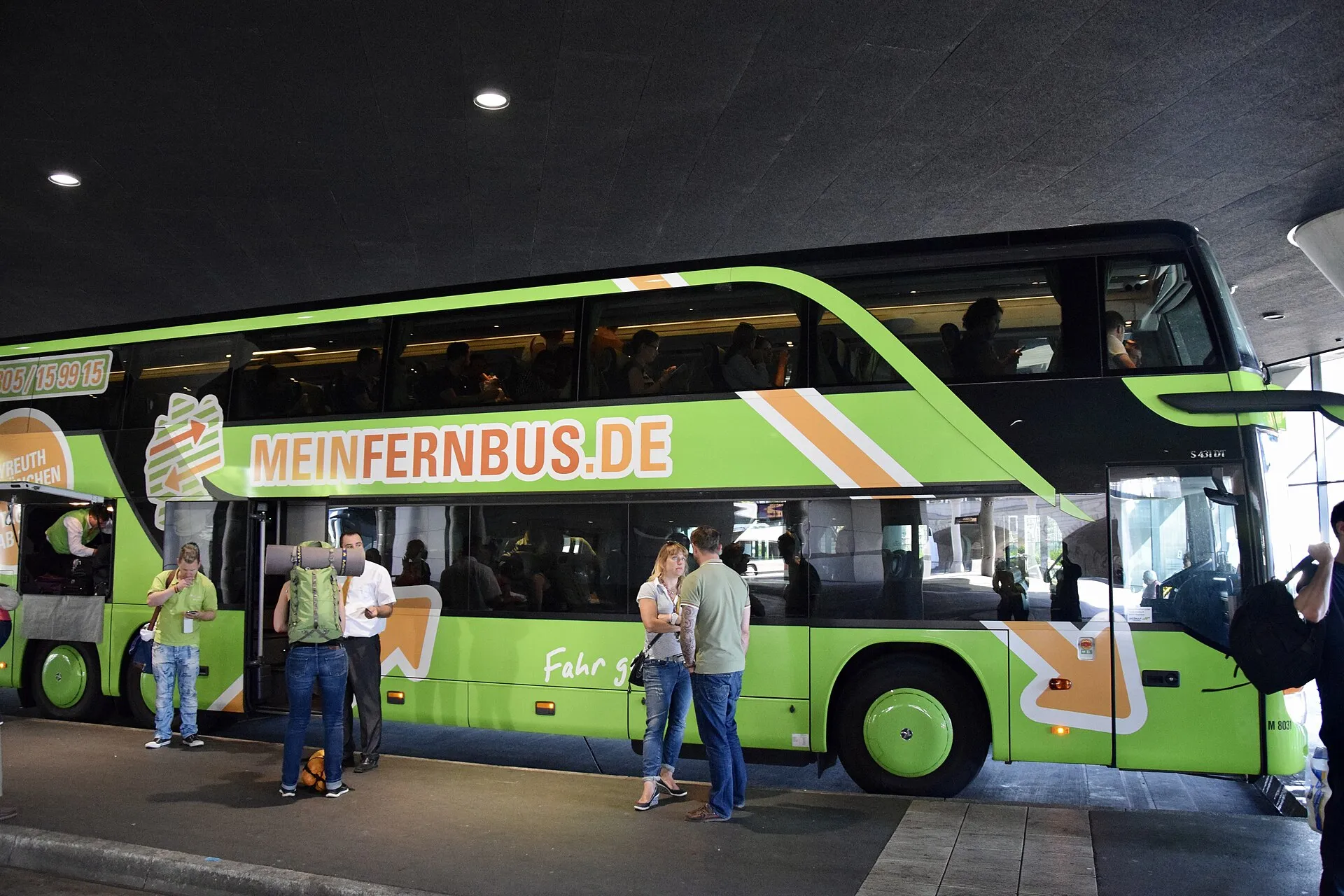 Yolcuların bagajlarıyla bindiği yeşil MEINFERNBUS.DE/FlixBus çift katlı otobüsü, kalkış öncesi otobüs durağında bekleyen yolcular