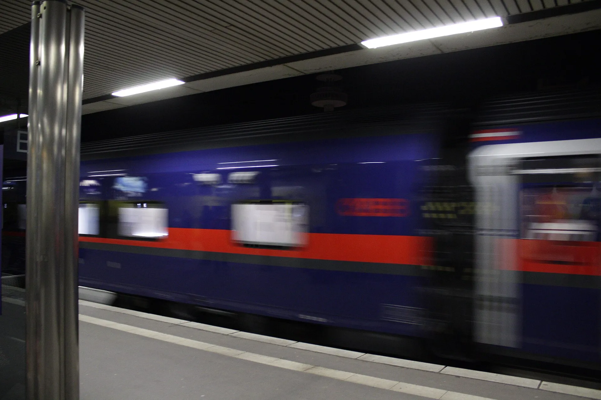 Bir istasyon peronundan hareket halinde geçen lacivert-kırmızı renkli ÖBB Nightjet gece treni vagonu, hareket bulanıklığıyla hızı yansıtıyor