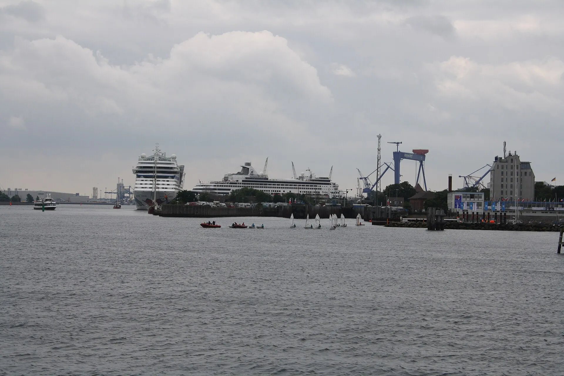 Rostock-Warnemünde limanında küçük yelkenli optimist tekneleri ve şişme botlar, arka planda demirli kruvaziyer gemileriyle birlikte görülüyor