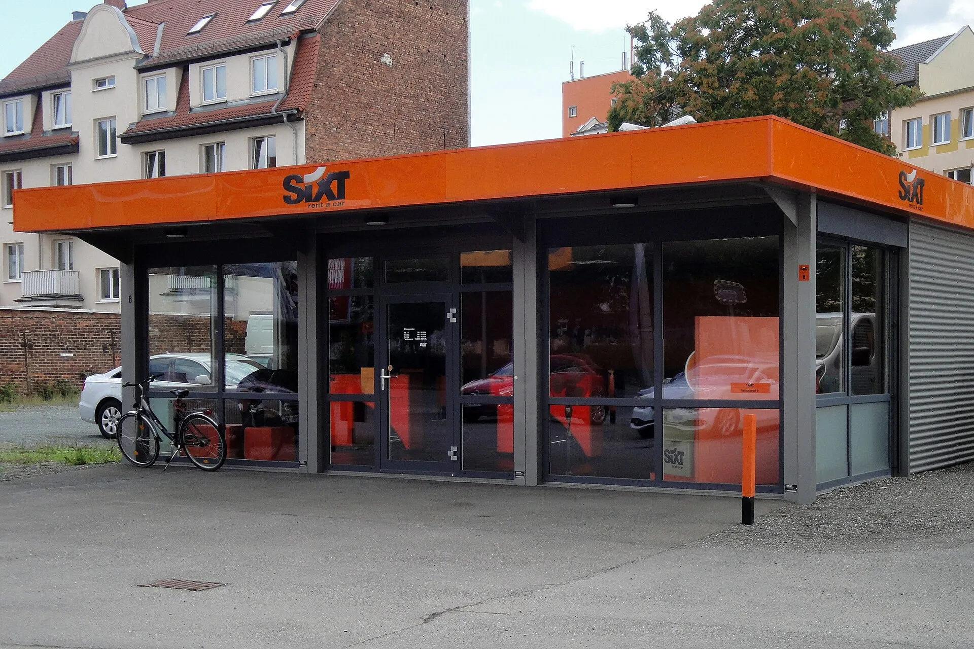 Almanya'da bir sokak kenarında turuncu tabelalı Sixt araç kiralama şubesi binası ve önünde park halindeki kiralık otomobiller